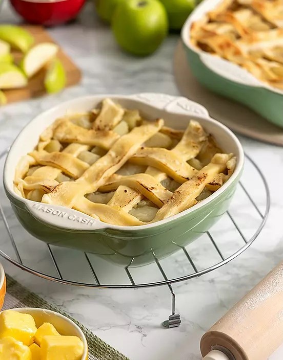 Mason Cash Fruits Apple Baker in green, filled with an apple pie and displayed on a cooling rack, with fresh apples and baking ingredients in the background.